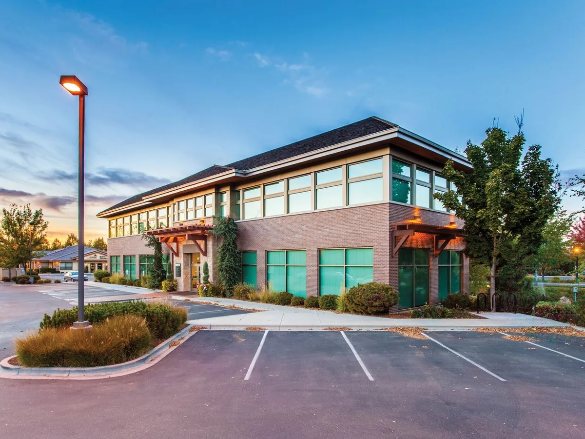 Low-rise brick-and-glass office building with mature trees, landscaped entry, and surface parking